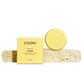 COOKI Shampoo Bar LEMON, featuring a square yellow box and a round yellow shampoo bar resting on a textured stone slab against a warm beige background.