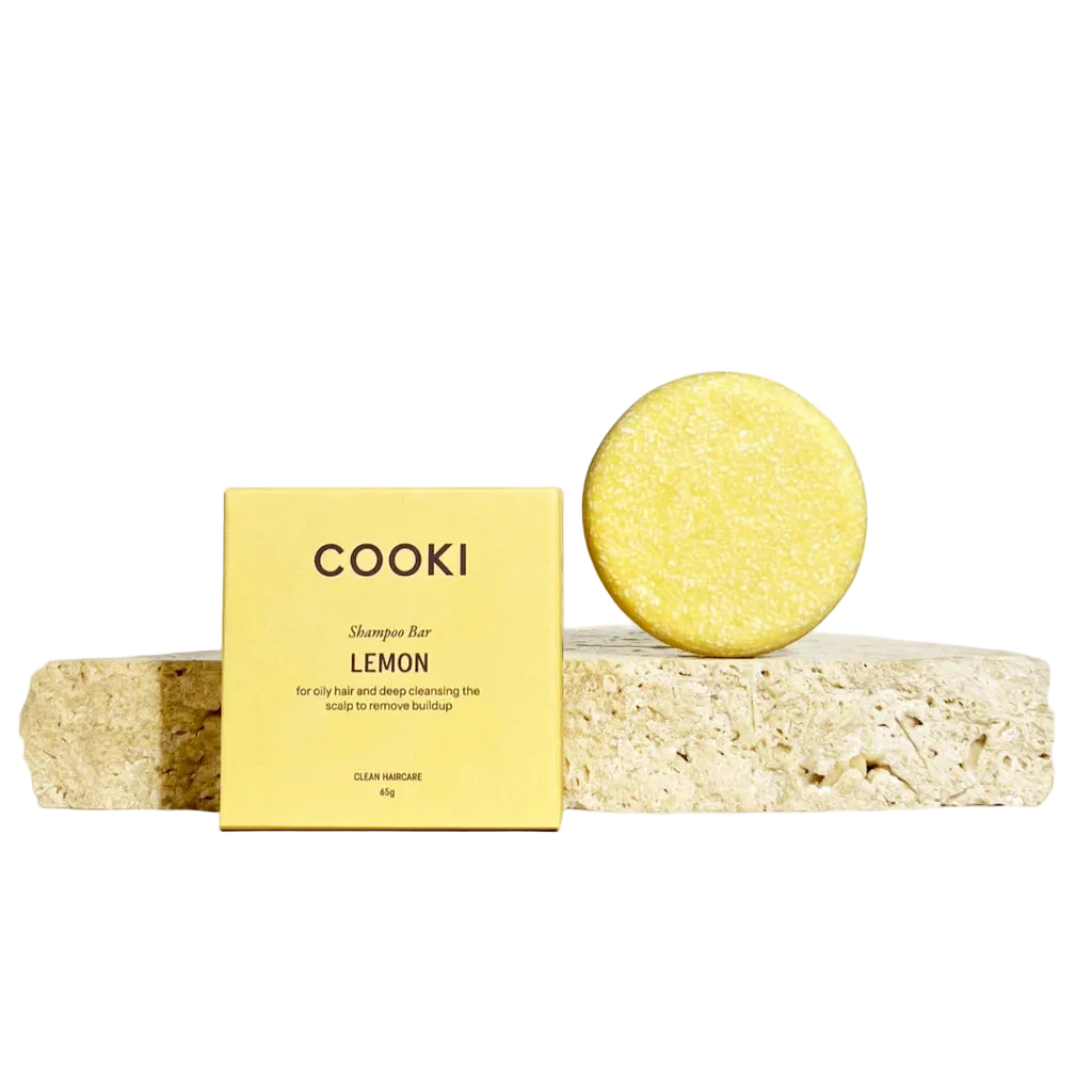 COOKI Shampoo Bar LEMON, featuring a square yellow box and a round yellow shampoo bar resting on a textured stone slab against a warm beige background.