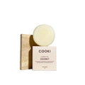 A round bar of COOKI Coconut Conditioner Bar standing upright on a beige stone block next to its white packaging box, set against a white background.