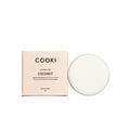 A round white shampoo bar next to its pale pink packaging box, labeled 'COOKI Shampoo Bar COCONUT', set on a textured beige surface with soft shadow overlays.
