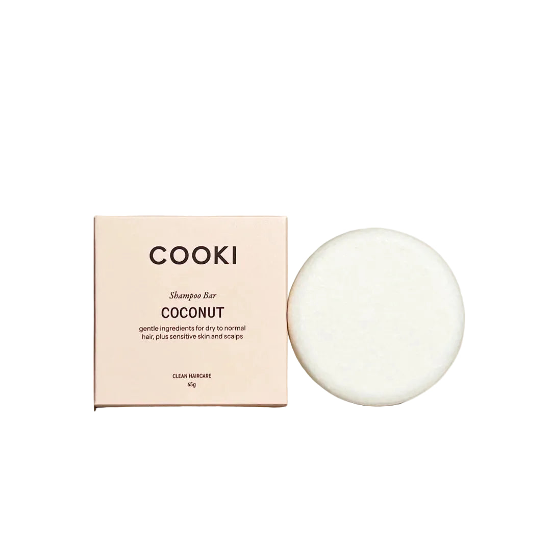 A round white shampoo bar next to its pale pink packaging box, labeled 'COOKI Shampoo Bar COCONUT', set on a textured beige surface with soft shadow overlays.