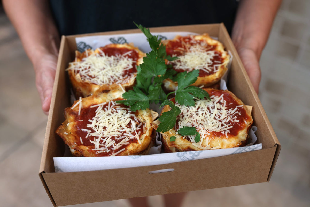 Four Greensmith Kitchen lasagnes in a takeaway box, topped with tomato sauce, grated cheese and fresh parsley.